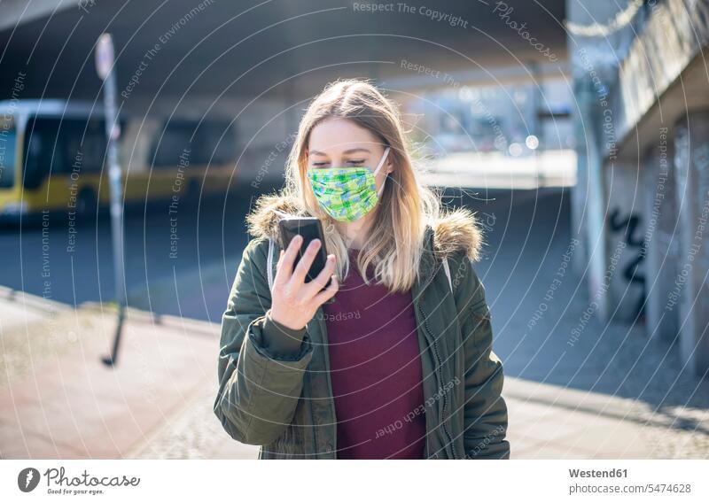 Junge Frau mit Smartphone mit Maske in der Stadt Informationen Jacken Transport Transportwesen KFZ Verkehrsmittel Autobus Autobusse Busse Omnibus Omnibusse