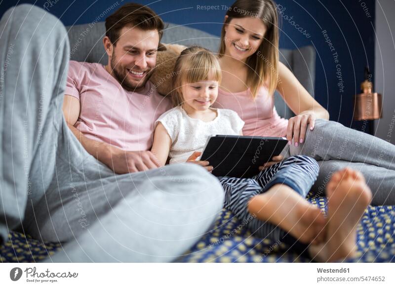 glückliche Familie sitzt auf dem Bett und schaut etwas auf dem digitalen Tablet mit der kleinen Tochter Familien Betten Töchter Glück glücklich sein