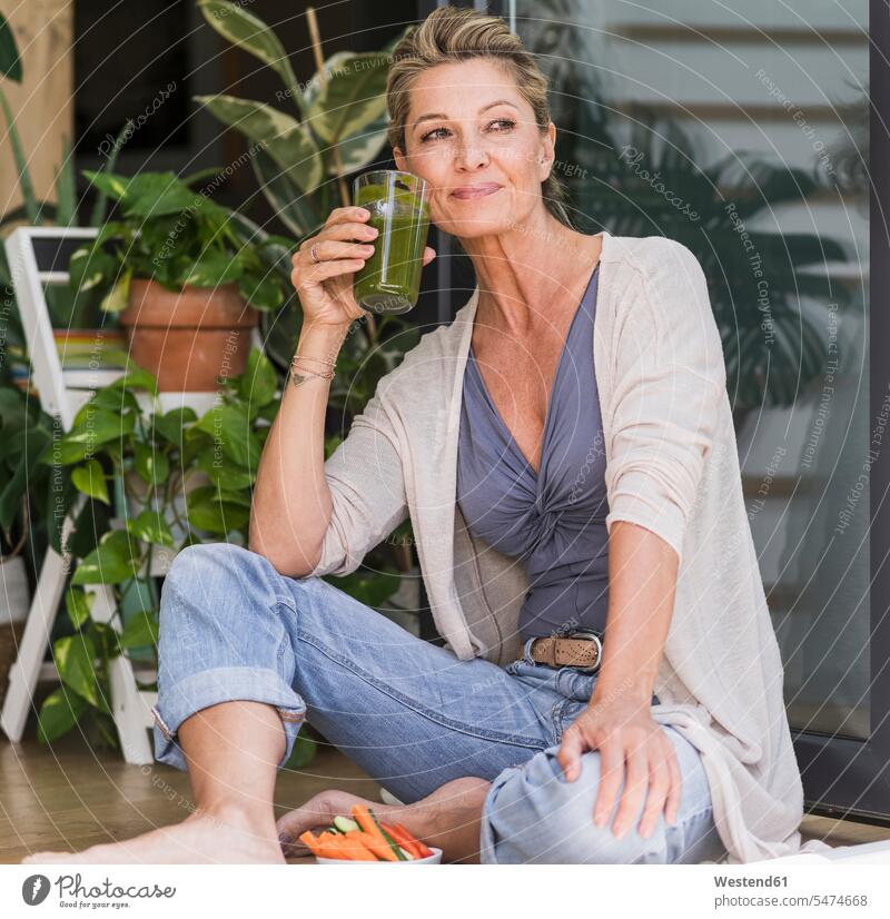 Porträt einer lächelnden reifen Frau, die an einer offenen Terrassentür sitzt und grünen Smoothie trinkt Gläser Trinkglas Trinkgläser Schale Schalen Schälchen