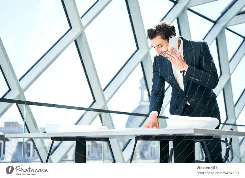 Lächelnder Geschäftsmann am Handy betrachtet Plan im Büro ansehen telefonieren anrufen Anruf telephonieren Pläne Businessmann Businessmänner Geschäftsmänner