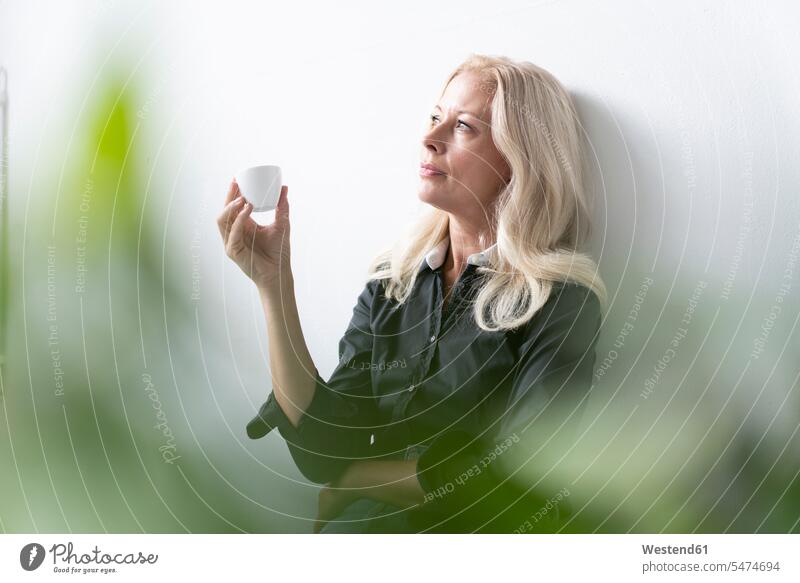 Nachdenkliche weibliche Fachkraft hält Kaffeetasse, während sie im Büro an einer weißen Wand sitzt Farbaufnahme Farbe Farbfoto Farbphoto Deutschland