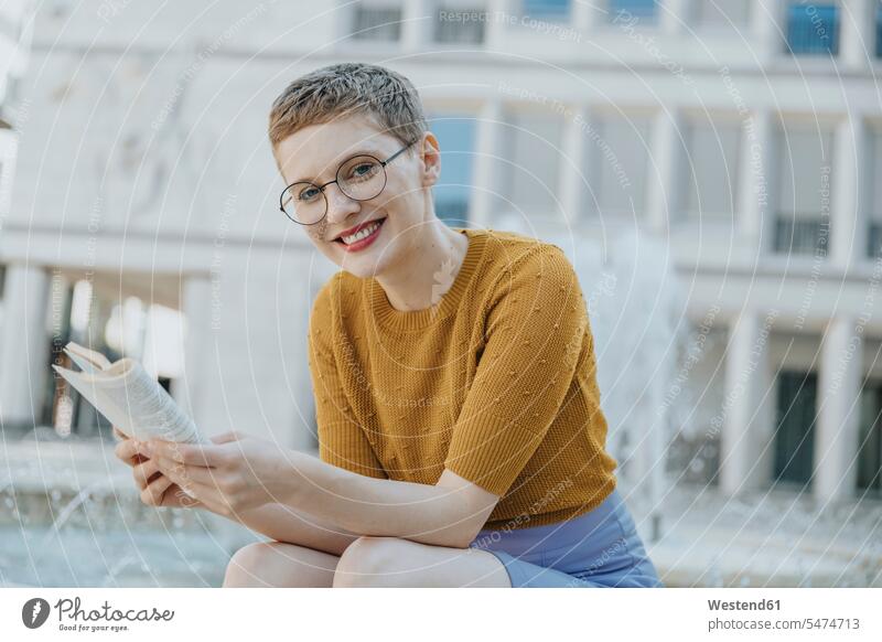 Lächelnde, mittelgroße erwachsene Frau mit Buch am Brunnen sitzend Farbaufnahme Farbe Farbfoto Farbphoto Außenaufnahme außen draußen im Freien Tag