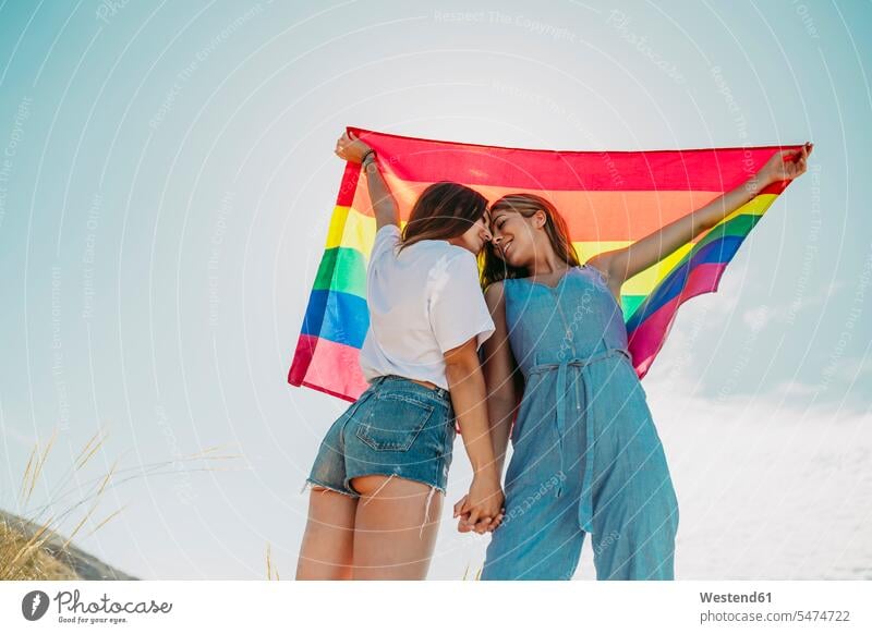 Zwei liebevolle junge Frauen mit einer LGBT-Flagge unter blauem Himmel (value=0) Freunde Kameradschaft Freundin Leute Menschen People Person Personen Paare