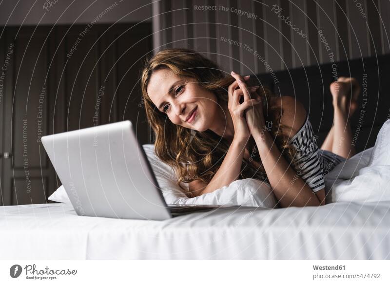 Lächelnde Frau, die zu Hause auf dem Bett liegt und auf den Laptop schaut Notebook Laptops Notebooks liegen liegend Betten weiblich Frauen ansehen Zuhause