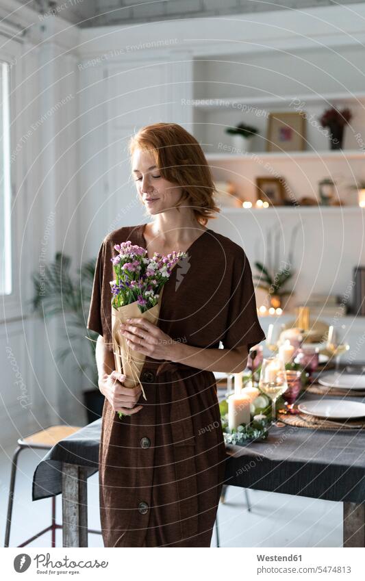 Rothaarige Frau, die einen Blumenstrauß hält, steht vor einem gedeckten Tisch Regal Ablage Regale Dreiviertelansicht Vorbereitung vorbereiten Überraschung