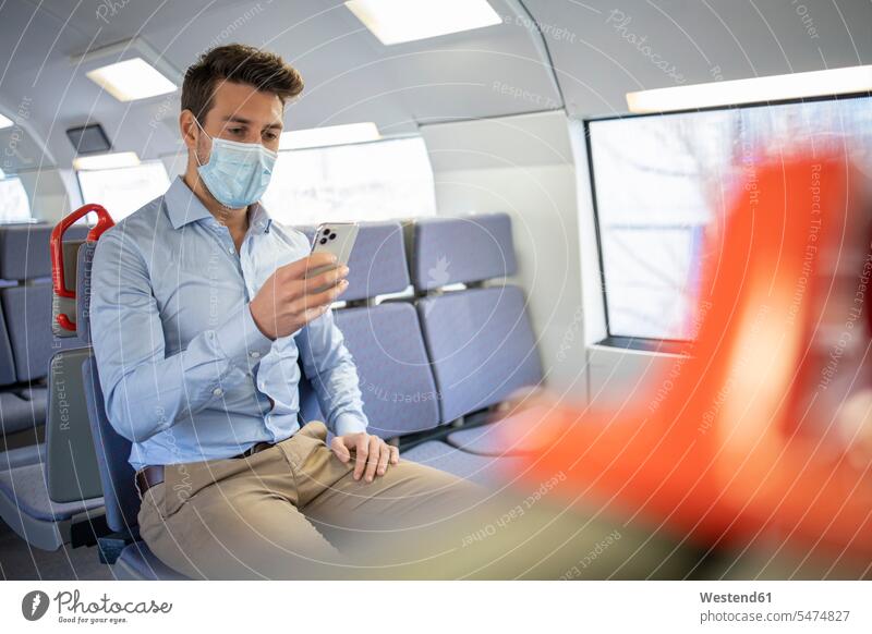 Geschäftsmann mit Gesichtsschutzmaske, der ein Mobiltelefon benutzt, während er im Zug sitzt Farbaufnahme Farbe Farbfoto Farbphoto Fahrzeuginnenraum Tag