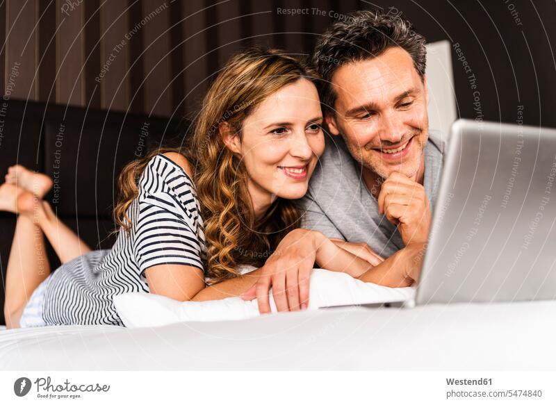 Glückliches Paar liegt zu Hause auf dem Bett und schaut auf den Laptop Pärchen Paare Partnerschaft liegen liegend Betten ansehen glücklich glücklich sein