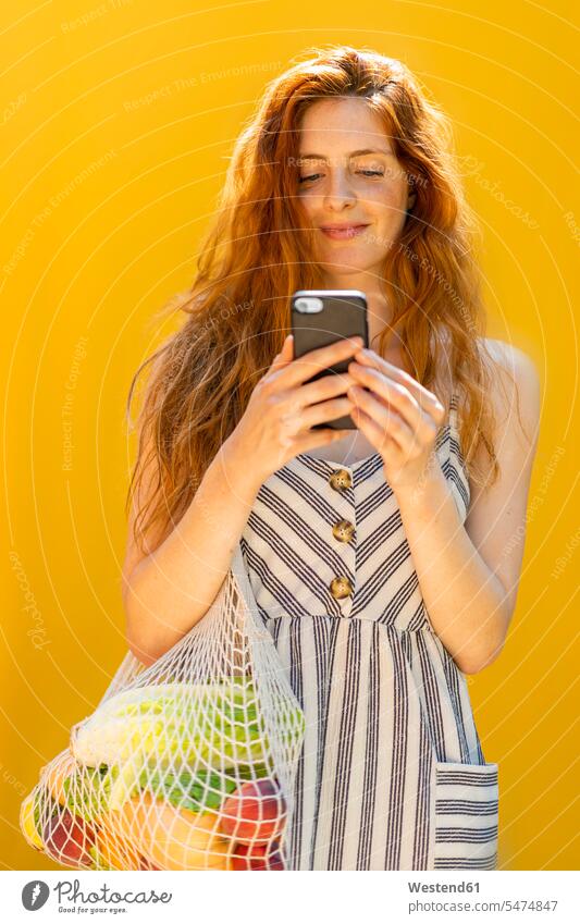 Lächelnde junge Frau, die ein Smartphone benutzt, während sie vor gelbem Hintergrund steht Farbaufnahme Farbe Farbfoto Farbphoto Spanien Freizeitbeschäftigung