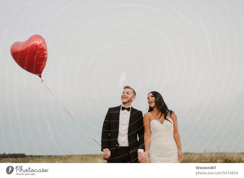 Glückliche Braut und Bräutigam mit herzförmigem Luftballon gegen den Himmel Farbaufnahme Farbe Farbfoto Farbphoto Außenaufnahme außen draußen im Freien Tag