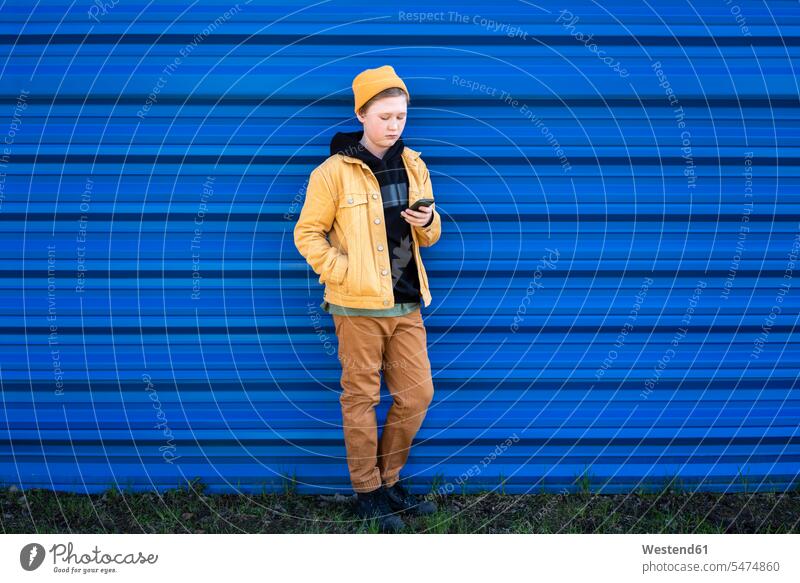 Junge benutzt Smartphone, während er gegen den blauen Verschluss steht Farbaufnahme Farbe Farbfoto Farbphoto Außenaufnahme außen draußen im Freien Tag