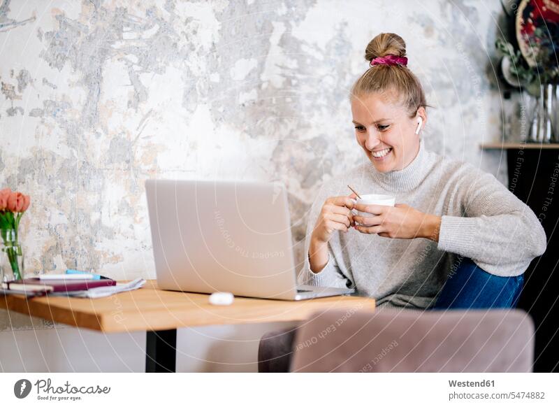 Lächelnde blonde Frau sitzt zu Hause auf einem Sessel und führt Video-Chat mit Laptop und Kopfhörern Leute Menschen People Person Personen Europäisch Kaukasier