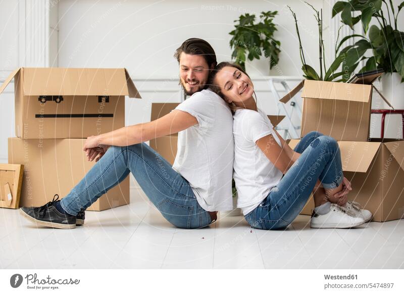 Glückliches Paar mit Kartons auf dem Boden sitzend im neuen Zuhause lächeln zu Hause daheim Umzug umziehen Pappkartons Wohnung Mietwohnungen wohnen Böden Boeden