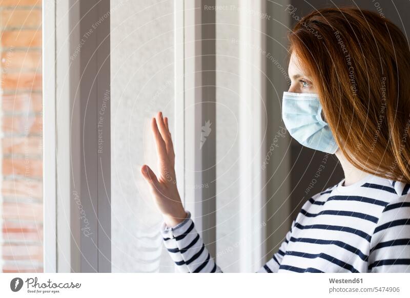 Rothaarige Frau mit Gesichtsschutzmaske, die während COVID-19 am Fenster ihres Hauses steht Farbaufnahme Farbe Farbfoto Farbphoto Innenaufnahme Innenaufnahmen