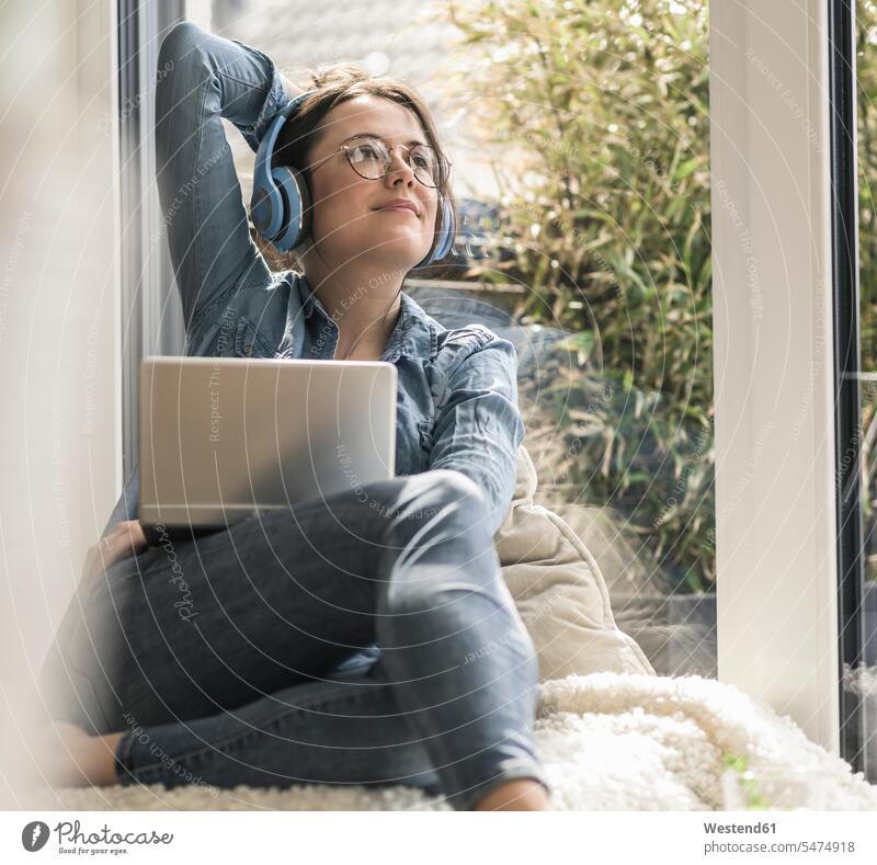 Frau mit Kopfhörer und Laptop sitzt am Fenster zu Hause Kopfhoerer Zuhause daheim weiblich Frauen lächeln Notebook Laptops Notebooks sitzen sitzend Erwachsener