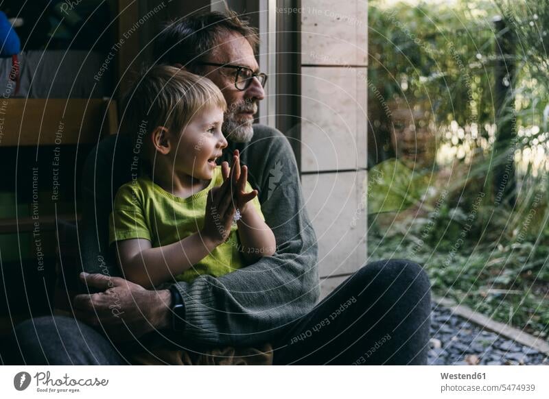 Vater und kleiner Sohn sitzen zu Hause zusammen auf dem Boden und schauen aus dem Fenster Leute Menschen People Person Personen Familien Familienvater Papa Papi