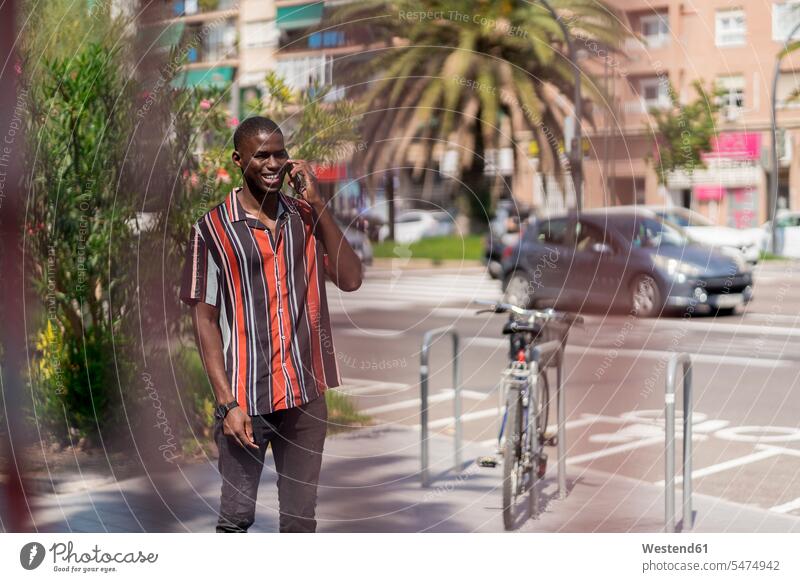 Lächelnder junger Mann spricht mit einem Smartphone, während er auf einem Fußweg in der Stadt steht Farbaufnahme Farbe Farbfoto Farbphoto Tag Tageslichtaufnahme