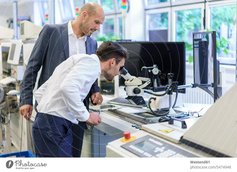 Junger Ingenieur schaut durch ein Mikroskop, während er einem Geschäftsmann im Labor zur Seite steht Farbaufnahme Farbe Farbfoto Farbphoto Innenaufnahme