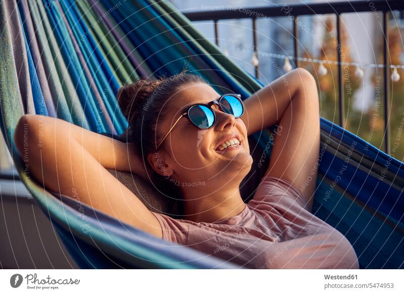 Porträt einer glücklichen jungen Frau mit Sonnenbrille, die auf einer Hängematte auf dem Balkon liegt Leute Menschen People Person Personen Europäisch Kaukasier