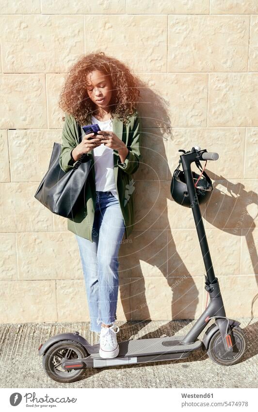 Junge Frau, die ein Smartphone benutzt, während sie mit einem elektrischen Roller auf dem Bürgersteig steht Farbaufnahme Farbe Farbfoto Farbphoto Außenaufnahme