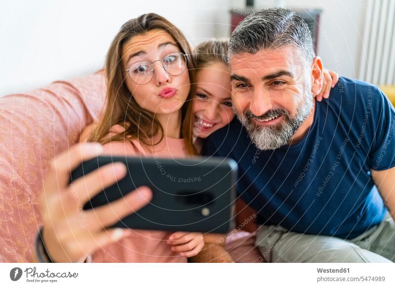 Glücklicher Vater mit zwei Töchtern nimmt Selfie auf Couch zu Hause T-Shirts Couches Liege Sofas Telekommunikation telefonieren Handies Handys Mobiltelefon