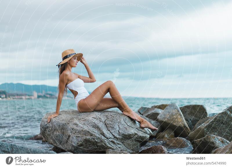 Schöne junge Frau trägt Badeanzug und Hut sitzt auf Felsen im Meer Meere sitzen sitzend weiblich Frauen schön Badeanzuege Badeanzüge Hüte Gewässer Wasser