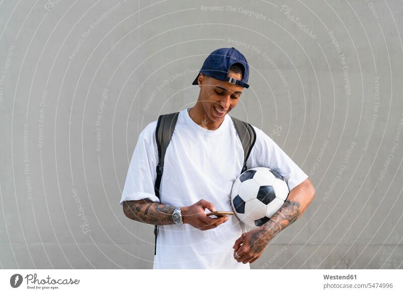 Tätowierter junger Mann mit Fussball schaut auf Smartphone Rucksäcke Informationen T-Shirts Bälle Fußbälle Telekommunikation Handies Handys Mobiltelefon