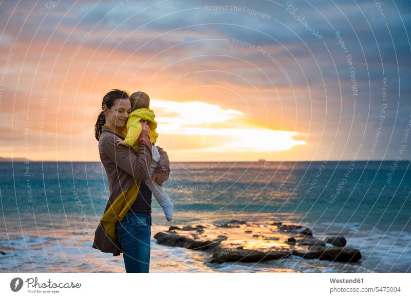 Mutter hält kleine Tochter bei Sonnenuntergang am Strand Mami Mutti Mütter Mama Töchter Baby Babies Babys Säuglinge Kind Kinder halten Sonnenuntergänge Beach