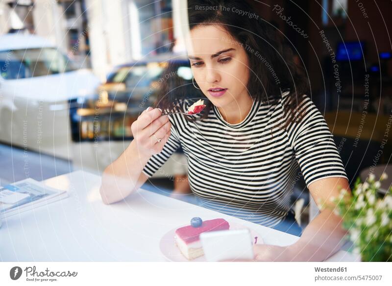 Junge Frau benutzt Handy und isst Kuchen in einem Café in der Stadt Mobiltelefon Handies Handys Mobiltelefone Cafe Kaffeehaus Bistro Cafes Cafés Kaffeehäuser