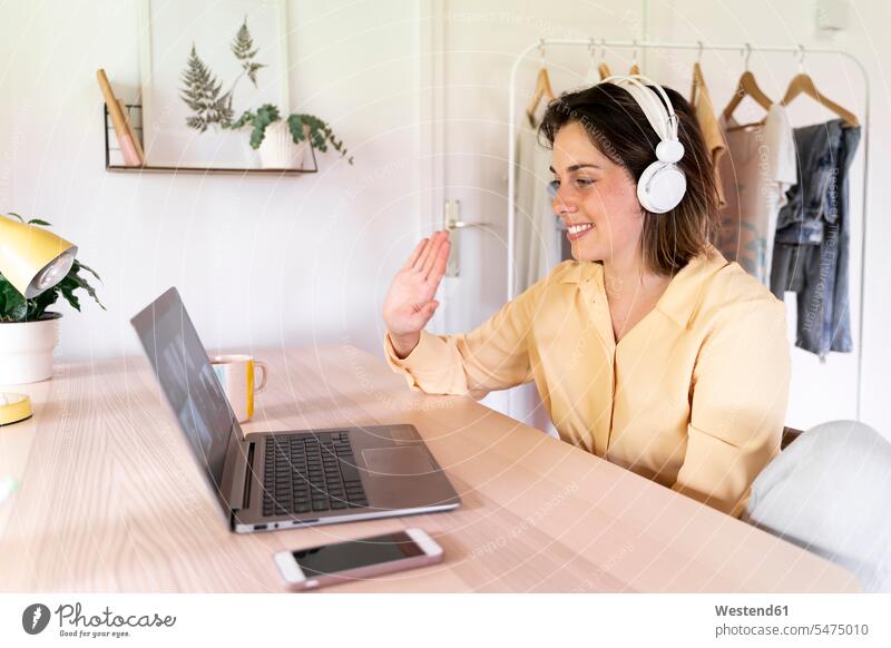 Lächelnde Frau winkt Freunden bei Videoanruf über Laptop zu Hause zu Farbaufnahme Farbe Farbfoto Farbphoto Spanien Innenaufnahme Innenaufnahmen innen drinnen