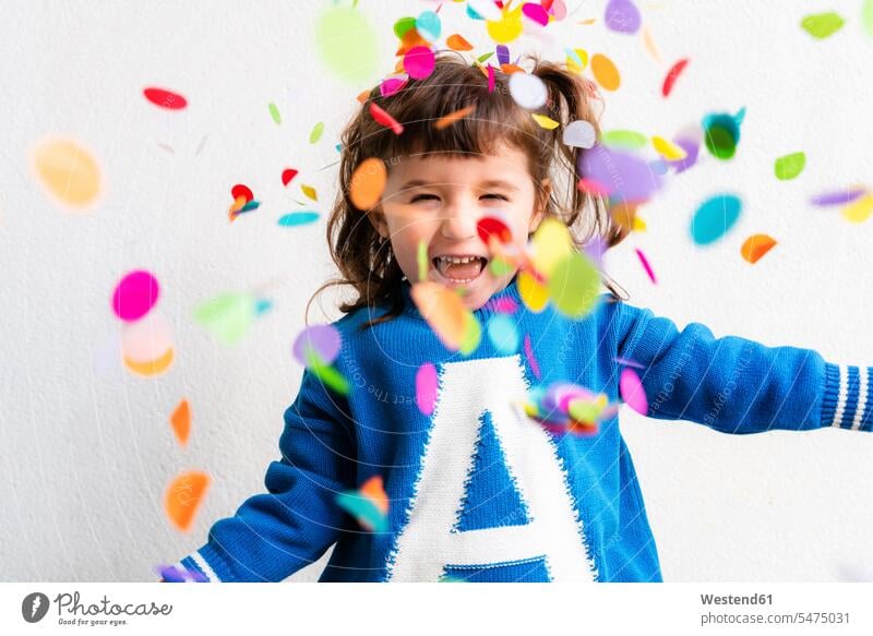 Glückliches kleines Mädchen, das auf einer Party vor einer weißen Wand Konfetti bläst Papiere Confetti abstürzen hinfallen stuerzen Sturz begeistert