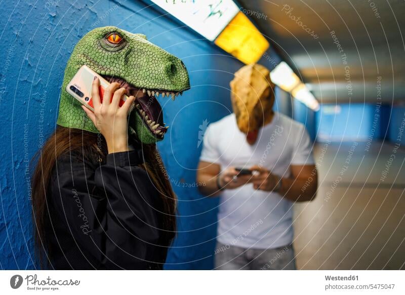 Frau telefoniert am Smartphone mit einem männlichen Freund, während sie eine Dinosaurier-Maske an der blauen Wand trägt Farbaufnahme Farbe Farbfoto Farbphoto