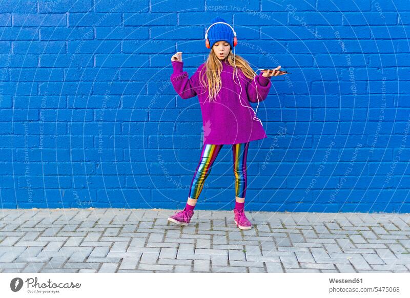 Mädchen mit blauer Mütze und übergroßem rosa Pullover hört Musik mit Kopfhörern vor einer blauen Wand Spanien Leggings Leggins Smartphone iPhone Smartphones