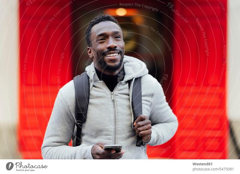 Lächelnder Mann mit Handy, der wegschaut, während er im Freien steht Farbaufnahme Farbe Farbfoto Farbphoto Außenaufnahme außen draußen Tag Tageslichtaufnahme