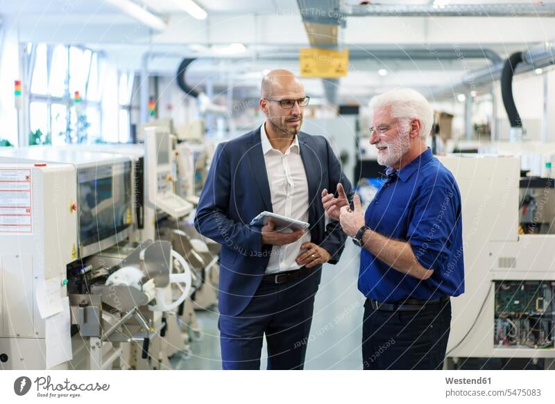 Geschäftsmann hält digitales Tablett in der Hand, während er einen leitenden Manager bei einer Diskussion in einer beleuchteten Fabrik beobachtet Farbaufnahme