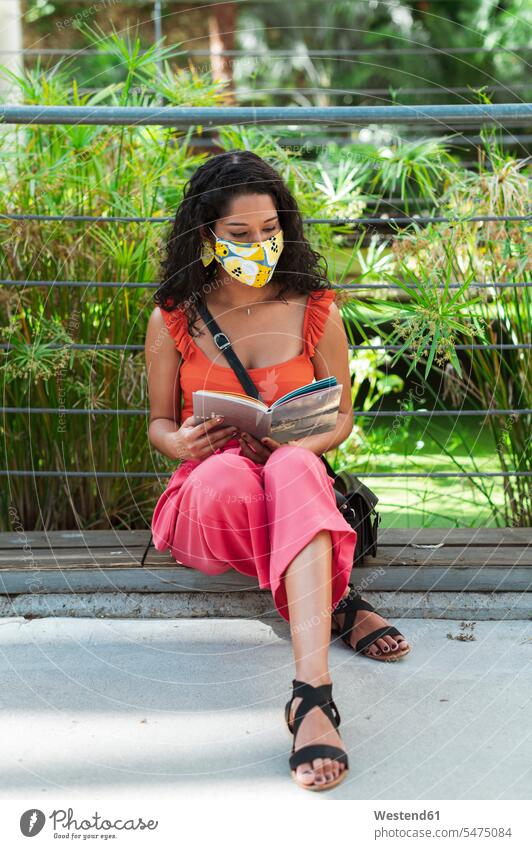 Junge Frau mit Schutzmaske beim Lesen eines Buches im Park sitzend während der COVID-19-Pandemie Farbaufnahme Farbe Farbfoto Farbphoto Außenaufnahme außen