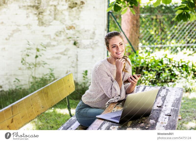Glückliche tagträumende Frau sitzt im Garten mit Laptop und Handy Tagtraum Gärten Gaerten Notebook Laptops Notebooks sitzen sitzend glücklich glücklich sein