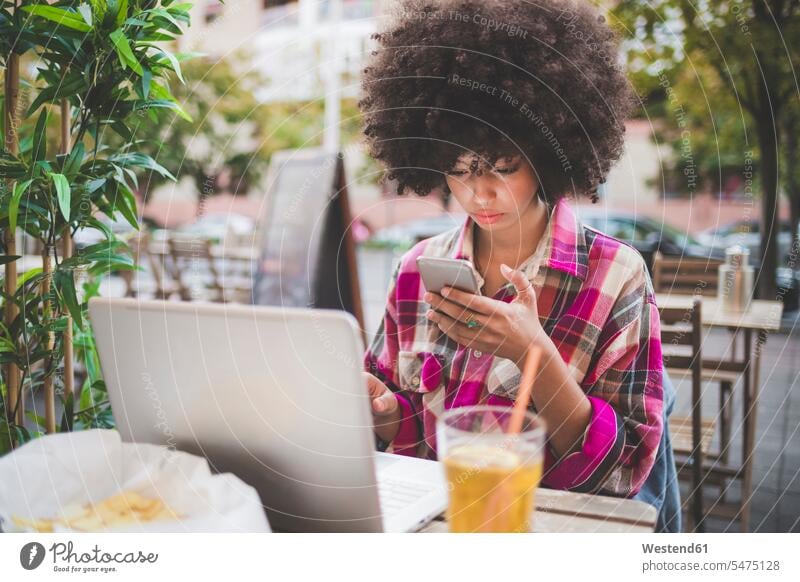 Junge Frau mit Afrofrisur mit Smartphone und Laptop in einem Straßencafé in der Stadt Leute Menschen People Person Personen gelockt gelockte Haare