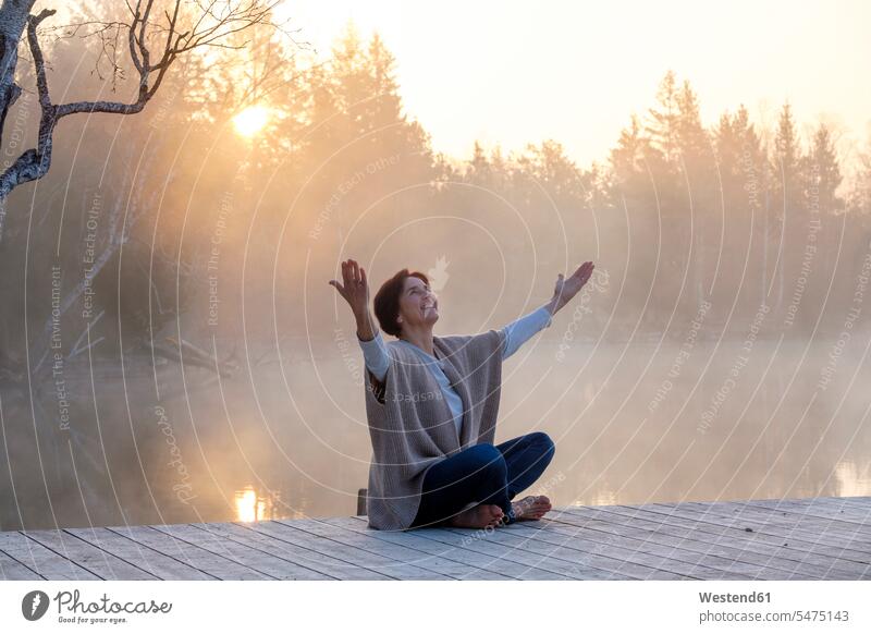 Erwachsene Frau meditiert bei nebligem Sonnenaufgang am Steg am Seeufer Außenaufnahme außen draußen im Freien Ufer Natur ländliches Motiv nicht städtisch Nebel