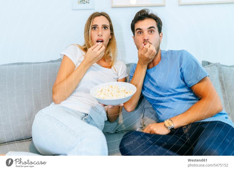 Aufgeregtes Paar sieht fern, während es zu Hause auf dem Sofa Popcorn isst Farbaufnahme Farbe Farbfoto Farbphoto Innenaufnahme Innenaufnahmen innen drinnen Tag
