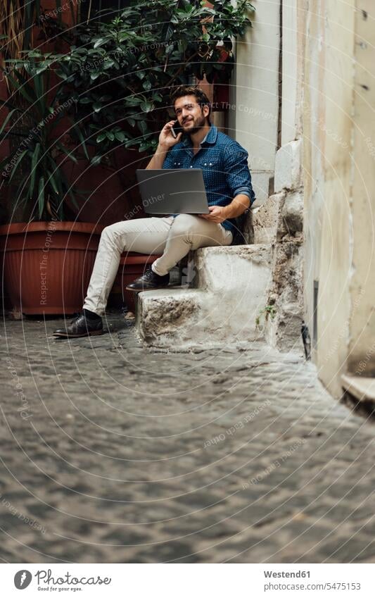 Lächelnde Geschäftsmann am Telefon sitzen auf Stufen in einem Hof mit Laptop Notebook Laptops Notebooks Innenhof telefonieren anrufen Anruf telephonieren Treppe