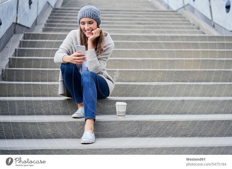 Glückliche Frau mit Kaffee, die auf der Treppe sitzt und auf ihr Handy schaut weiblich Frauen glücklich glücklich sein glücklichsein Treppenaufgang Coffee to go