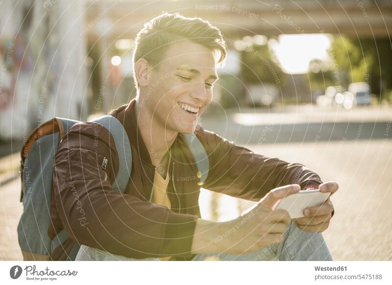 Glücklicher junger Mann, der ein Smartphone benutzt, während er an einem sonnigen Tag auf der Straße sitzt Farbaufnahme Farbe Farbfoto Farbphoto Außenaufnahme