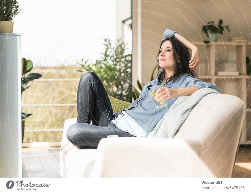 Lächelnde Frau mit einem Glas aufgegossenem Wasser auf der Couch zu Hause sitzend Zuhause daheim weiblich Frauen sitzt lächeln Sofa Couches Liege Sofas