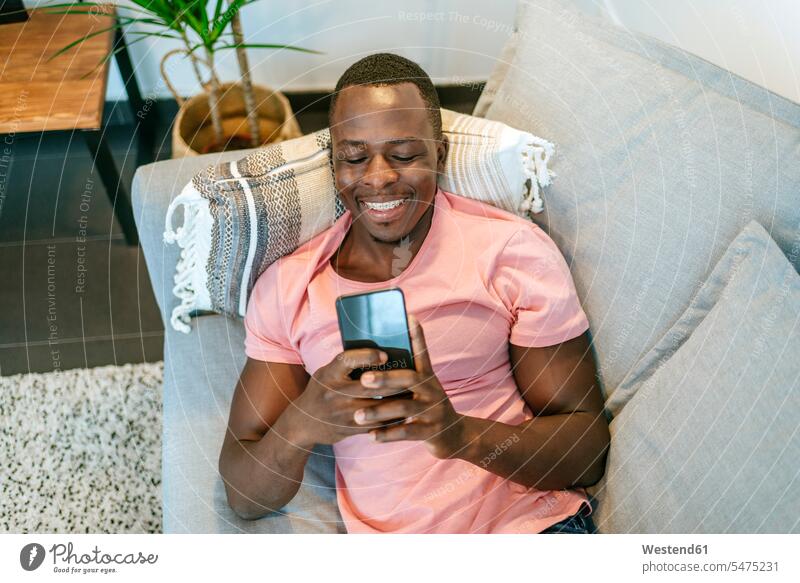 Glücklicher junger Mann auf der Couch liegend mit Smartphone zu Hause Leute Menschen People Person Personen Afrikanisch Afrikanische Abstammung dunkelhäutig