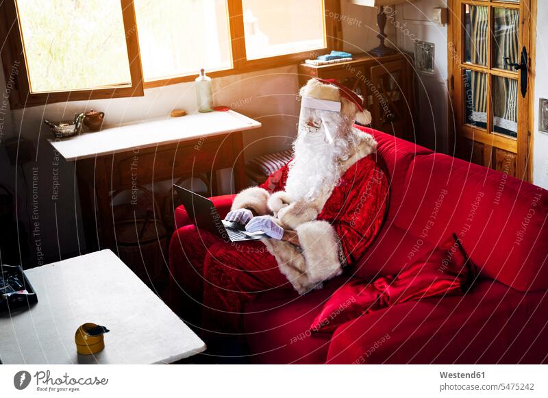 Mann trägt Weihnachtsmannkostüm mit Laptop, während er zu Hause auf dem Sofa sitzt Farbaufnahme Farbe Farbfoto Farbphoto Spanien Freizeitbeschäftigung Muße Zeit