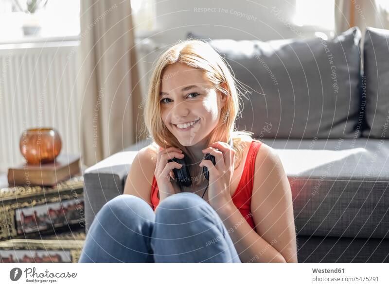Porträt einer glücklichen jungen Frau, die zu Hause auf dem Boden sitzt und mit Kopfhörern Musik hört Leute Menschen People Person Personen Alleinstehende