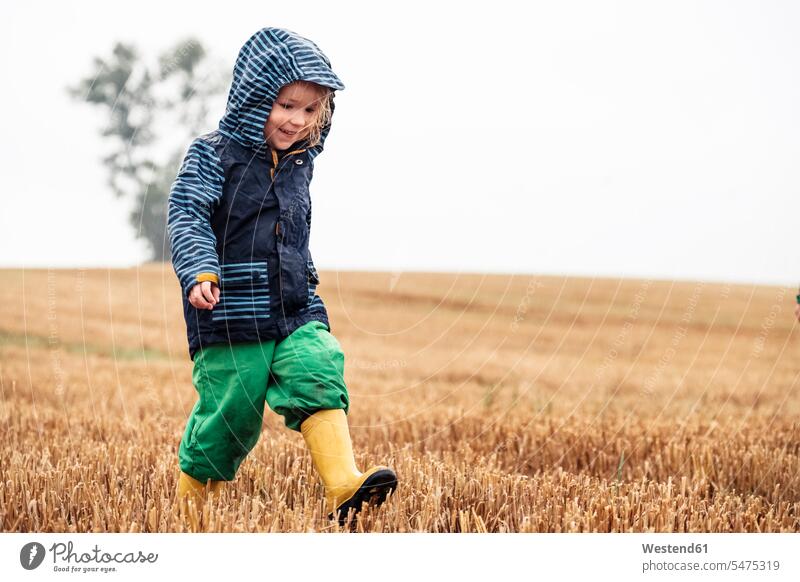 Glückliches kleines Mädchen geht an einem regnerischen Tag auf Stoppelfeld Kapuzen Schuhe begeistert Enthusiasmus enthusiastisch Überschwang Überschwenglichkeit