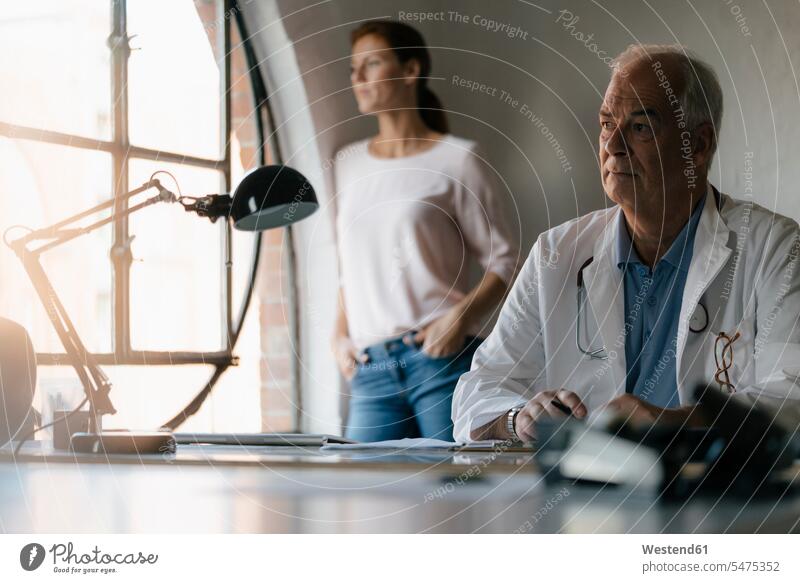 Seriöser Oberarzt sitzt am Schreibtisch in einer Arztpraxis mit Frau im Hintergrund ernst Ernst Ernsthaftigkeit ernsthaft Arztpraxen Praxis Senior ältere Männer
