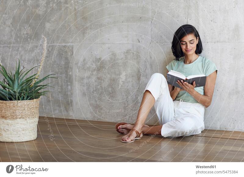 Geschäftsfrau liest Buch, während sie sich auf einem Parkettboden an der Wand im Büro entspannt Farbaufnahme Farbe Farbfoto Farbphoto Deutschland Innenaufnahme