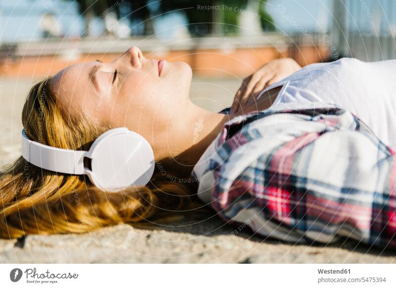 Auf dem Rücken liegende Frau, die an einem sonnigen Tag über Kopfhörer Musik hört Farbaufnahme Farbe Farbfoto Farbphoto Außenaufnahme außen draußen im Freien